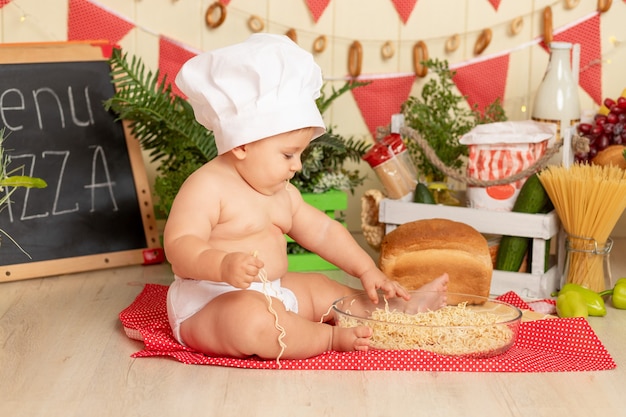 Little child cook in a hat sitting in the kitchen among the products and eating spaghetti