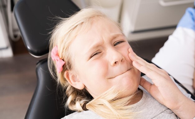 Маленький ребенок в кабинете дантиста в удобном кресле трогает рот рукой с болезненным выражением лица из-за зубной боли или болезни зубов. Изображение концепции стоматолога