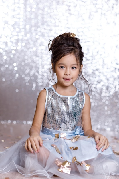 Photo little child girl in blue dress sitting on the floor with confetti on background with silver bokeh. birtday party