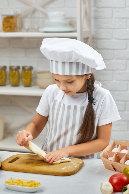 Маленькая девочка в кепке и фартуке месит тесто