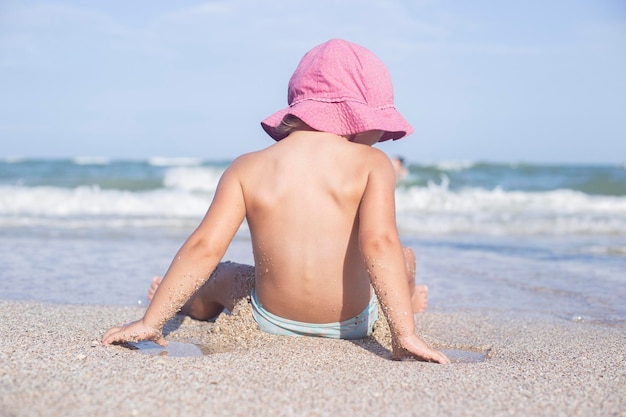 Little child girl in a hat sitting with her back on the beach.