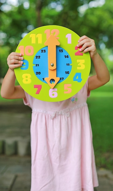 Foto ragazza del piccolo bambino che mostra l'orologio del giocattolo rotondo di 6 in punto nel giardino