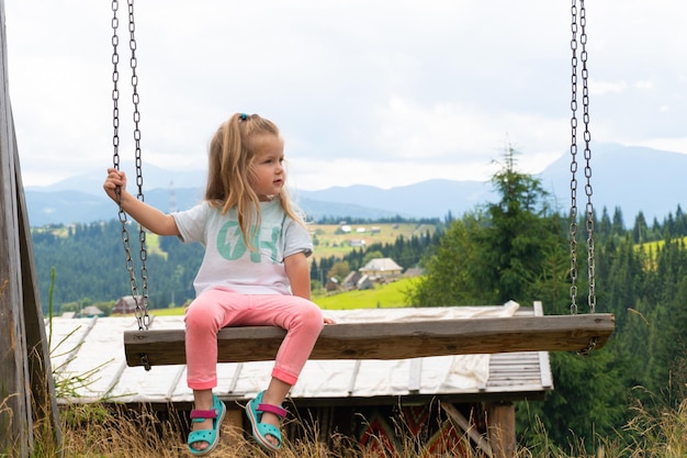 Little child girl sitting on swing with mountains behind. Outdoor countryside view