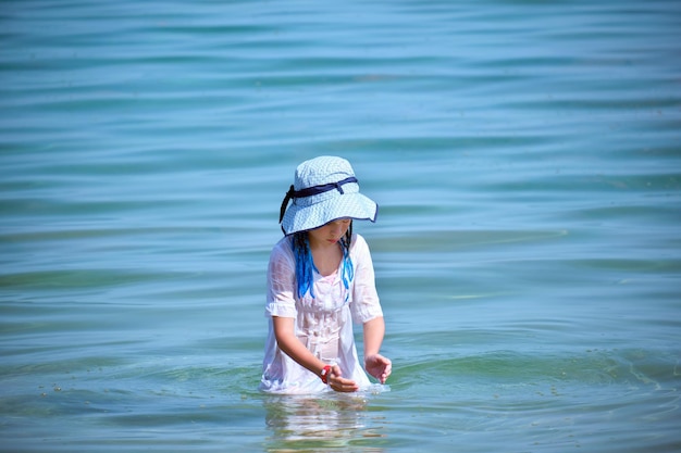 Bambina in abito bianco bagnato e grande cappello che gioca da sola nell'acqua di mare godendo le vacanze estive