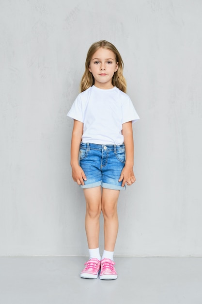 Little child girl in white t-shirt, jeans shorts and pink sneakers standing straight near the wall