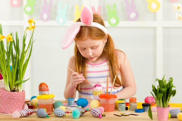 Маленькая девочка с ушами пасхального кролика рисует пасхальные яйца