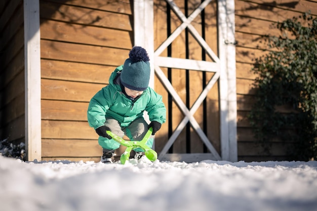 緑のジャケットとニットの帽子をかぶった小さな子供が家の近くで雪玉を作ります