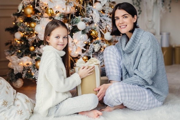 Little child and her mother sit on warm white carpet near decorated New Year tree holds present in hands have pleased and happy expressions glad to spend holidays together Childhood and holidays