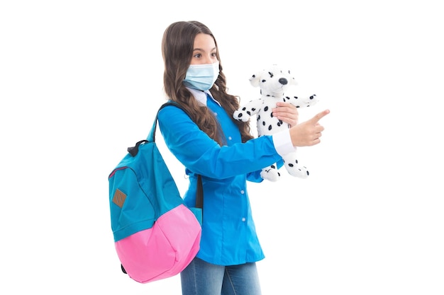 Little child in protective medical mask back to school with toy dog during COVID19 pandemic isolated on white pointing finger copy space