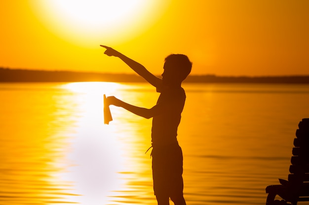 A little child stands on the lake at sunset. In the left hand he holds a paper airplane, and with the right hand he points with his finger into the distance