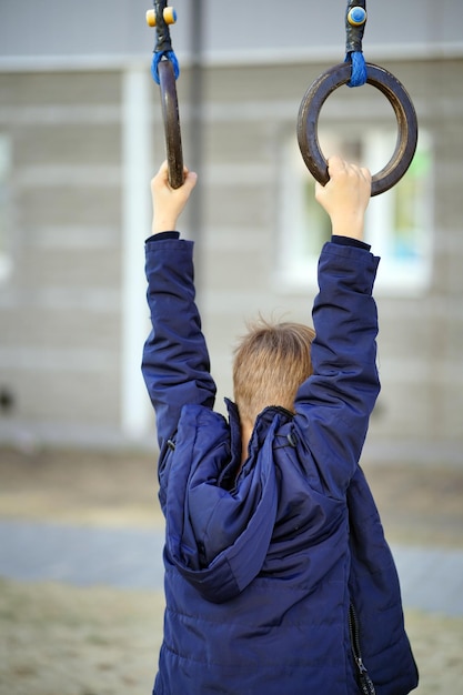 Little child swings on sports rings in yard of house Active boy playing on playground