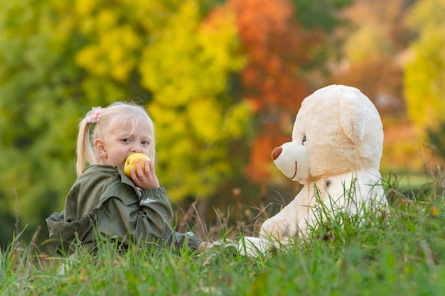 Little child on walk on autumn day in nature Blonde girl eats apple and plays with large teddy bear sitting on grass