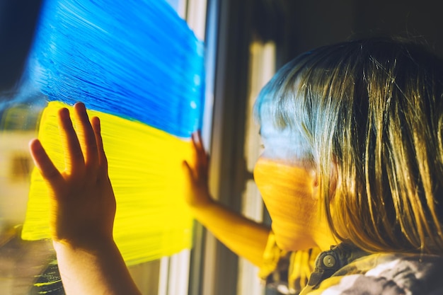 Little child with hands on yellowblue flag image of Ukraine on window Support Ukrainians Concept emotions Color symbol image of flag of Ukraine on glass with paints