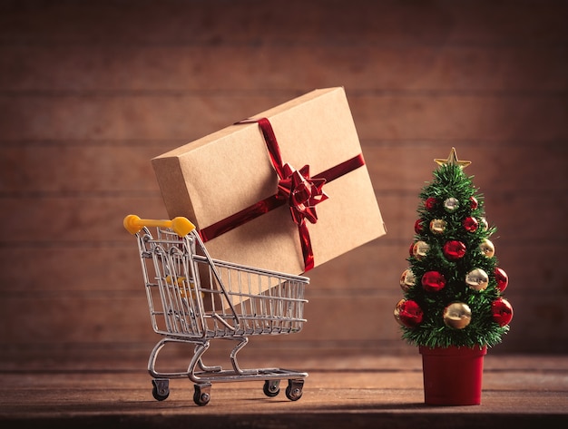 Little Christmas tree and gift box in supermarket cart on wooden table and background