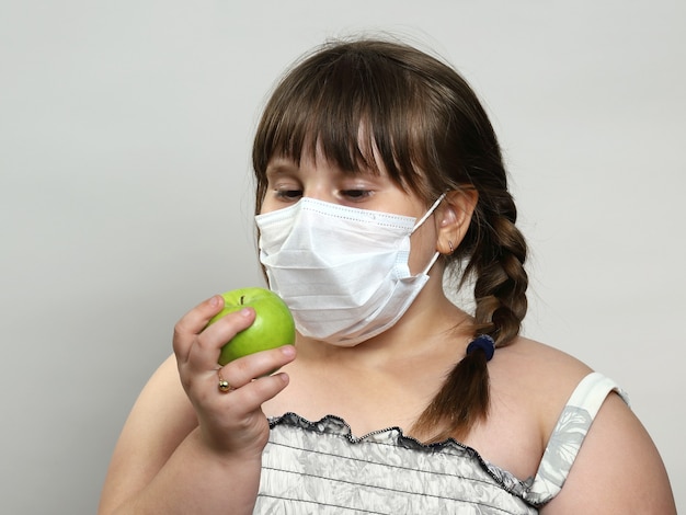 Little chubby girl in medical face mask holds green apple