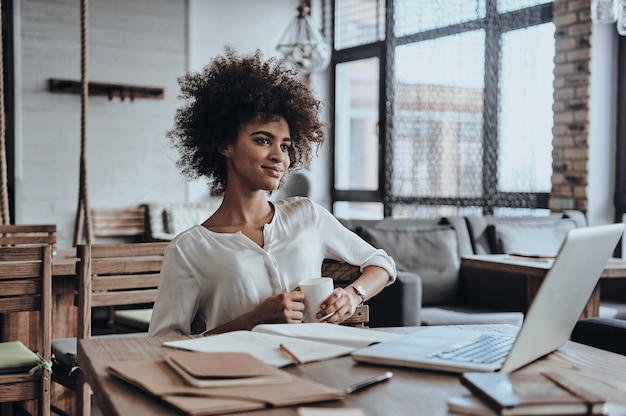 Little coffee break. Attractive young African woman holding a cup and looking away with smile 