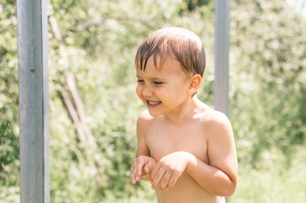 Little contented wet four year old kid boy with water drops on his skin after rinsing with cold water in a outdoor shower in nature in a garden in the village