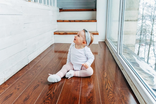 Little crawling baby girl one year old siting on floor in bright light living room near window smiling and laughing