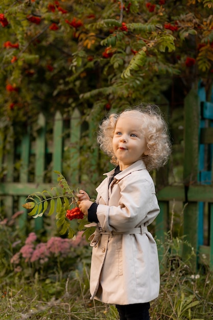 Little curly blonde girl with different emotions on her face in white raincoat is standing in grass