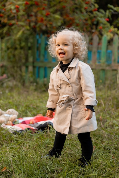 Little curly blonde girl with different emotions on her face in white raincoat is standing in grass