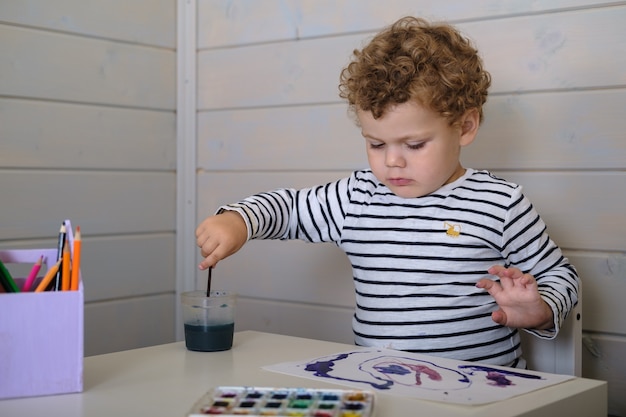 Little curly boy draws watercolor paints on paper sitting at the table