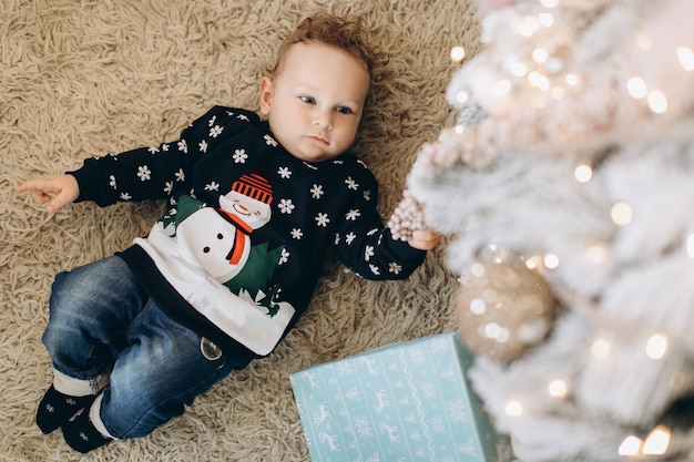 A little curly boy in a traditional christmas sweater spends\
time at home near the christmas tree