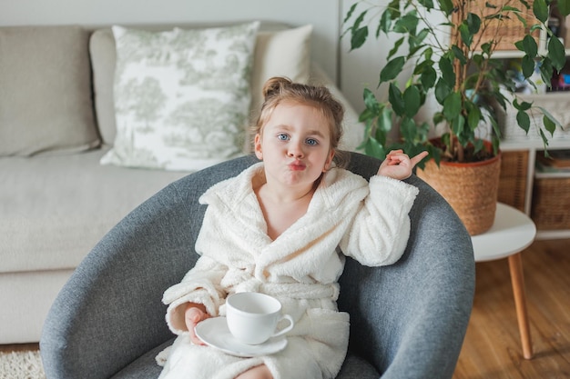 Little curlyhaired girl in a soft light dressing gown drinks\
tea and relaxes at home sitting in a comfortable chair the baby\
imitates her mother beautiful homely cozy interior