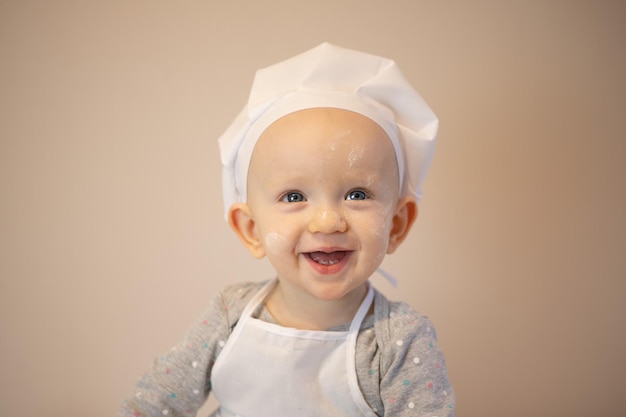 앞치마를 입은 작고 귀여운 아기와 주방장의 모자는 베이지색 배경에 고립되어 미소 짓는다
