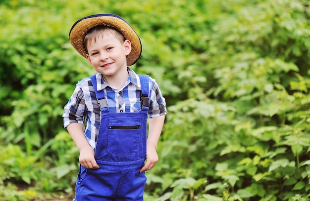 Piccolo neonato sveglio in cappello di paglia, camicia di plaid e tuta blu del lavoro su fondo verde.