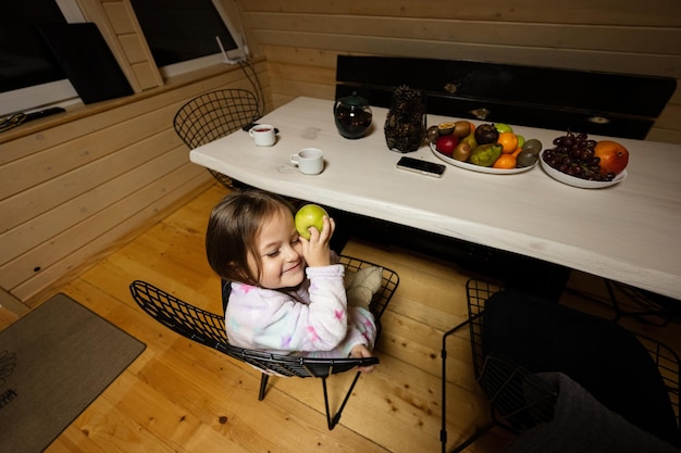 Little cute baby girl in pajama sit kitchen with green apple in cozy wooden tiny cabin house Life in countryside