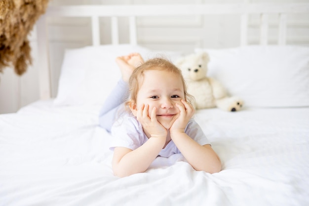 A little cute baby girl on a white cotton bed at home is indulging and having fun smiling on the bed at home with her hands folded under her cheeks