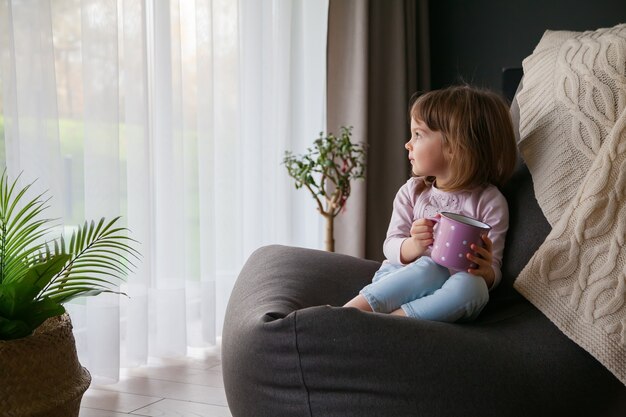 Little cute baby girl with a cup of tea looking through the window sitting on a cozy bean bag chair