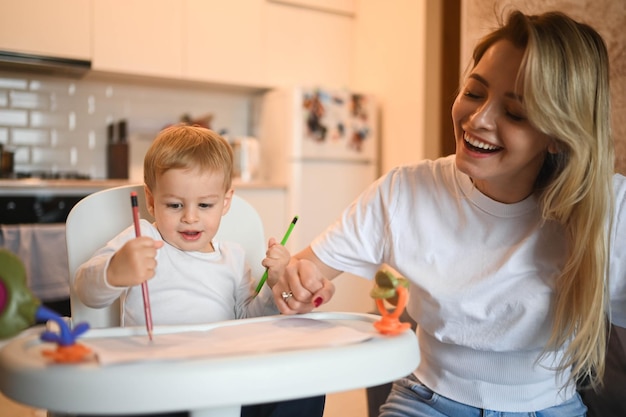 Piccolo bambino carino bambino biondo seduto sulla sedia del bambino che impara a disegnare una bella giovane mamma e