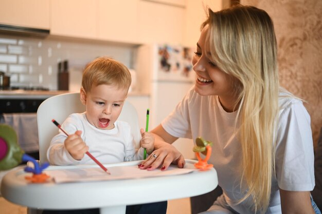 Piccolo bambino carino bambino biondo seduto sul seggiolone che impara a disegnare una bella giovane mamma e