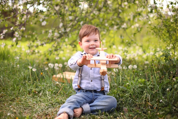 Маленький милый белокурый мальчик играет с деревянным самолетом в парке летом на траве в солнечный день, фокус на ребенка
