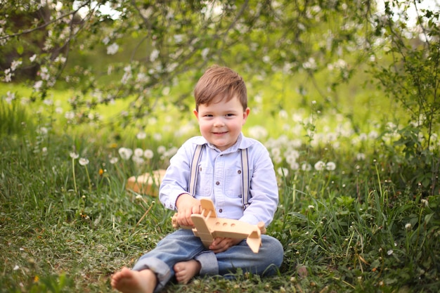 Little cute blond boy playing with a wooden plane in the summer park on the grass on a sunny day