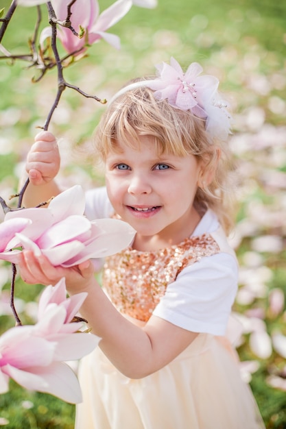 3歳の小さなかわいいブロンドの女の子は、開花するマグノリアの近くの公園で遊んでいます。春。