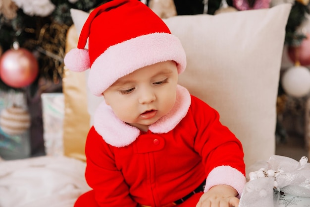 Little cute boy 6 months old dressed as Santa Claus sits near the Christmas tree. The baby sits on the floor with gifts near the Christmas tree. Christmas 2022