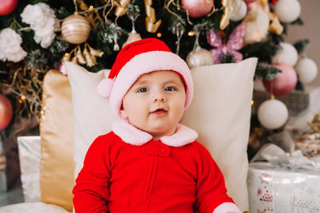 Little cute boy 6 months old dressed as Santa Claus sits near the Christmas tree. The baby sits on the floor with gifts near the Christmas tree. Christmas 2022