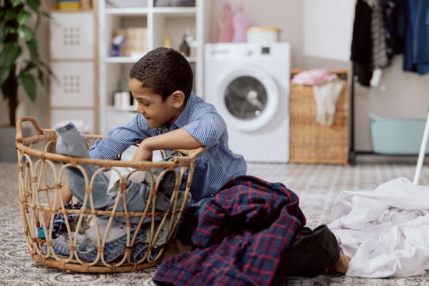 Little cute boy helps mom with housework sits on the floor in\
the bathroom laundry room takes clothes out of the hamper sorts\
before putting in the washing machine