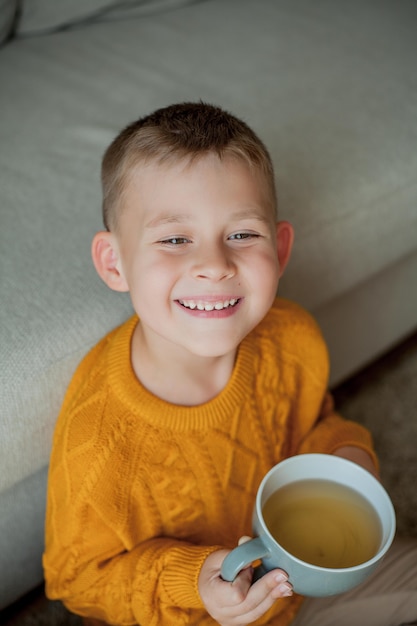 オレンジ色のセーターを着た小さなかわいい男の子がお茶を飲んでいます。家に座っている男の子の居心地の良い肖像画。秋。