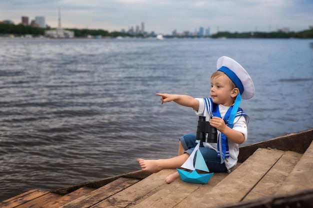 船乗りの帽子をかぶった小さなかわいい男の子が海岸に座って双眼鏡をのぞき、ボートで遊ぶ