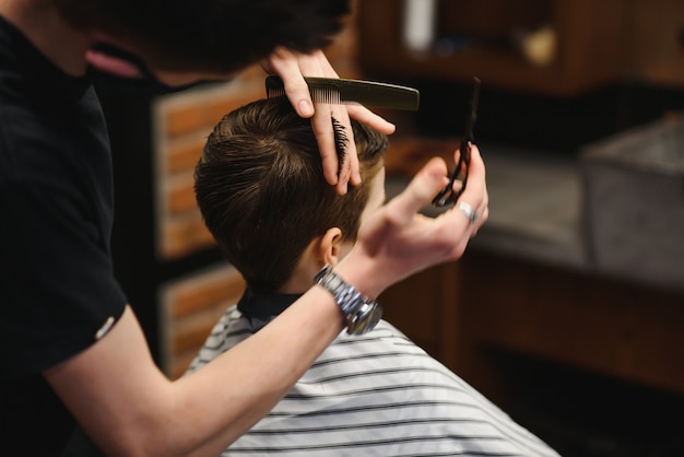 Un ragazzino carino siede in un parrucchiere dallo stilista, uno scolaro si fa tagliare i capelli in un salone di bellezza, un bambino da un barbiere, un taglio di capelli da uomo corto.