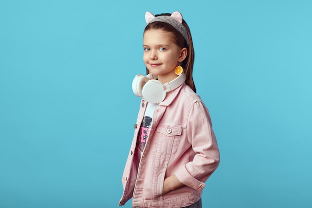 Little cute caucasian girl in pink jacket holding white headphones on neck