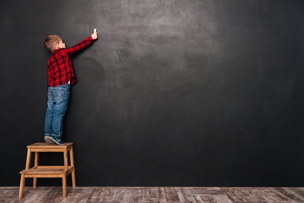 Little cute child standing on stool near blackboard and drawing on it