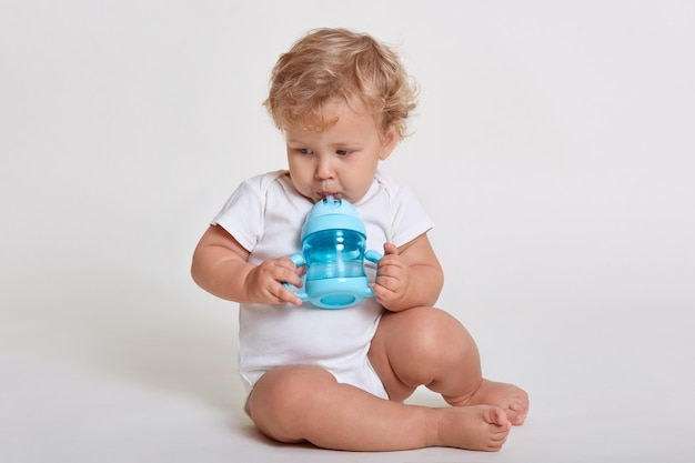 Little cute concentrated kid looking away while drinking water from baby cup, child with blonde curly hair sitting on floor