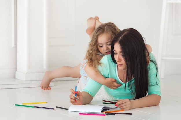 Маленькая милая дочь и ее мать, рисование красочными карандашами и с удовольствием вместе. милый ребенок и мама играя внутри помещения. счастливая семья, тратить время на рисование.