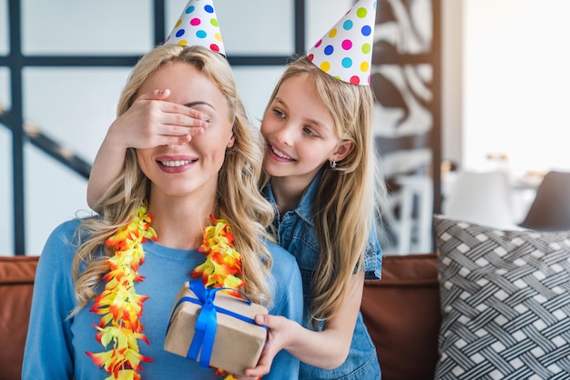 Little cute daughter closing eyes of excited mommy holding gift box on couch at home interior