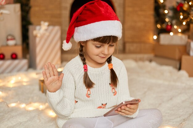 Little cute female kid with pigtails congratulation from home, smiling little girl using smart phone for video call, talks to somebody