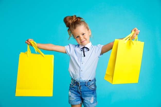 Little cute and funny girl holding big bright colored yellow paper bags in the blue studio.